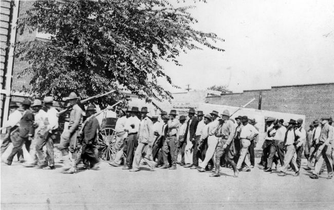 Um grupo de tropas da Guarda Nacional, carregando fuzis com baionetas presas, escolta homens negros desarmados para um centro de detenção após o Massacre de Tulsa, Tulsa, Oklahoma, em junho de 1921.
