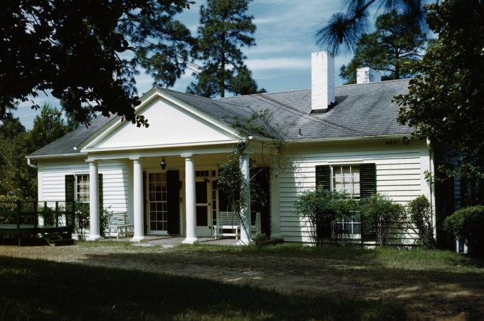 pequena casa branca com persianas pretas, amplo pórtico central com quatro colunas e frontão