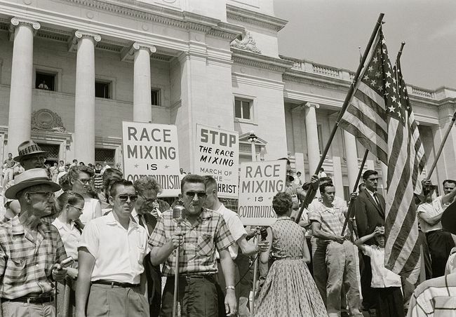 Pessoas segurando cartazes e bandeiras americanas protestando contra a admissão de " Little Rock Nine" na Central High School.