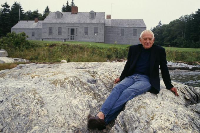 homem de cabelos brancos, sentado nas pedras em frente a uma casa na Nova Inglaterra