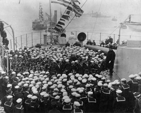 Presidente Theodore Roosevelt em pé em uma torre de navio de guerra com uma multidão de marinheiros à sua frente.