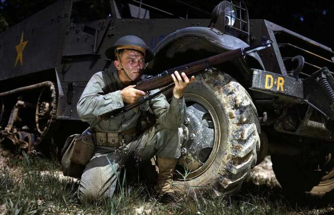 Um soldado americano se ajoelha com um M1 Garand na frente de um meio caminho.